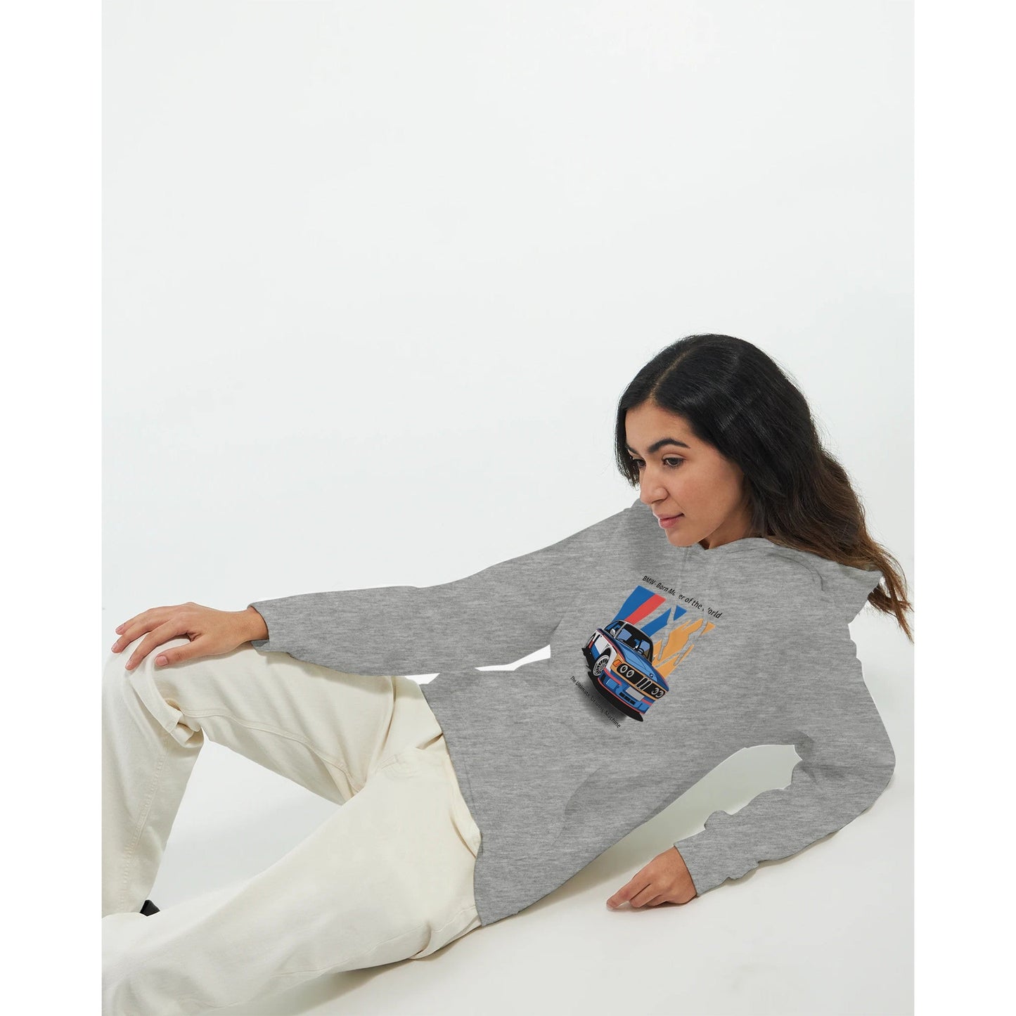 woman wearing a grey born to race unisex hoodie with a colorful car graphic, paired with white pants, lying down against a plain white background.