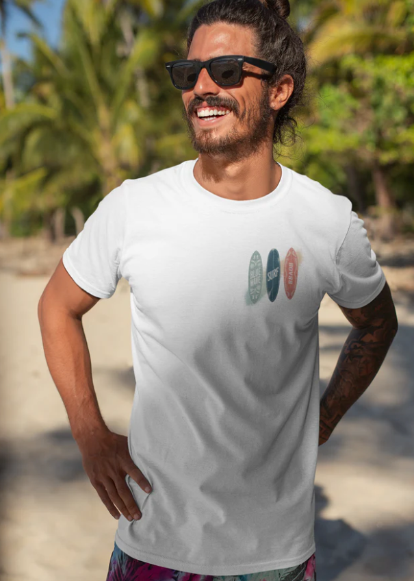 mann trägt weißes premium surfer t-shirt mit surfer-motiv am strand bei sonnenschein, lächelnd und entspannt.