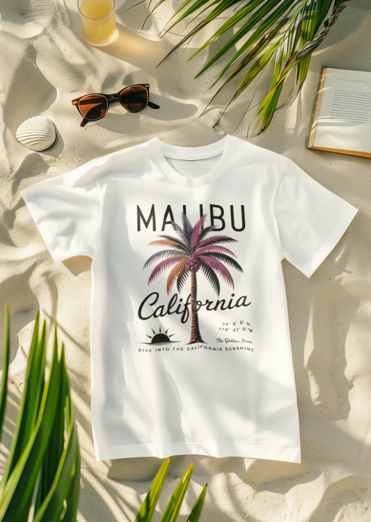 Weißes California Sunshine Sommer T-Shirt mit Malibu Palmenmotiv auf Sandstrand, umgeben von Sonnenbrille, Buch und Pflanze.