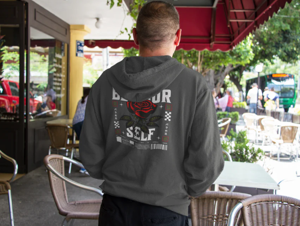 man wearing dark gray unisex hoodie with "be yourself" graphic and red rose design on back in outdoor café setting