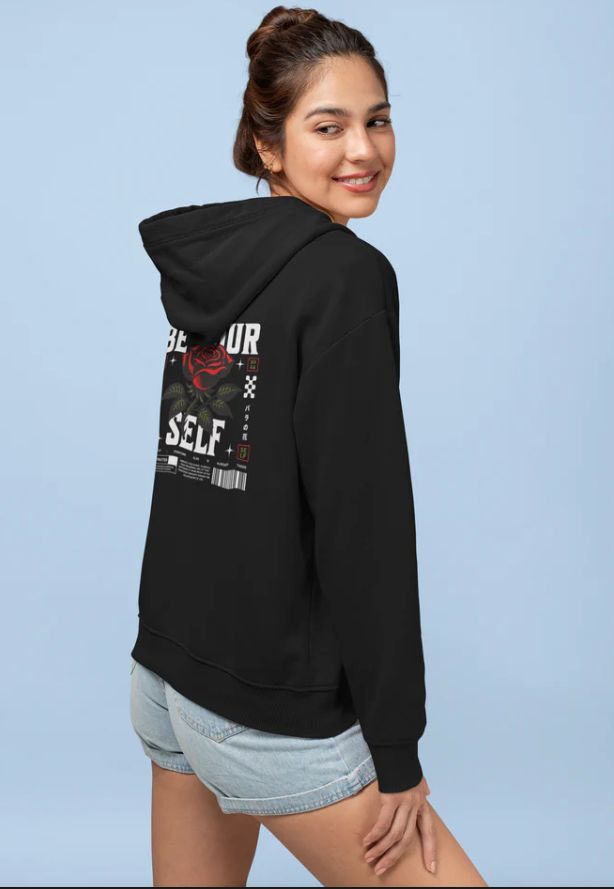 Black unisex hoodie with "Be Yourself" graphic design on back, worn by smiling woman with casual shorts.