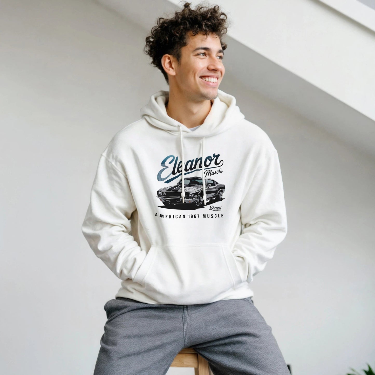young man wearing white eleanor hoodie with vintage muscle car print, sitting indoors and smiling